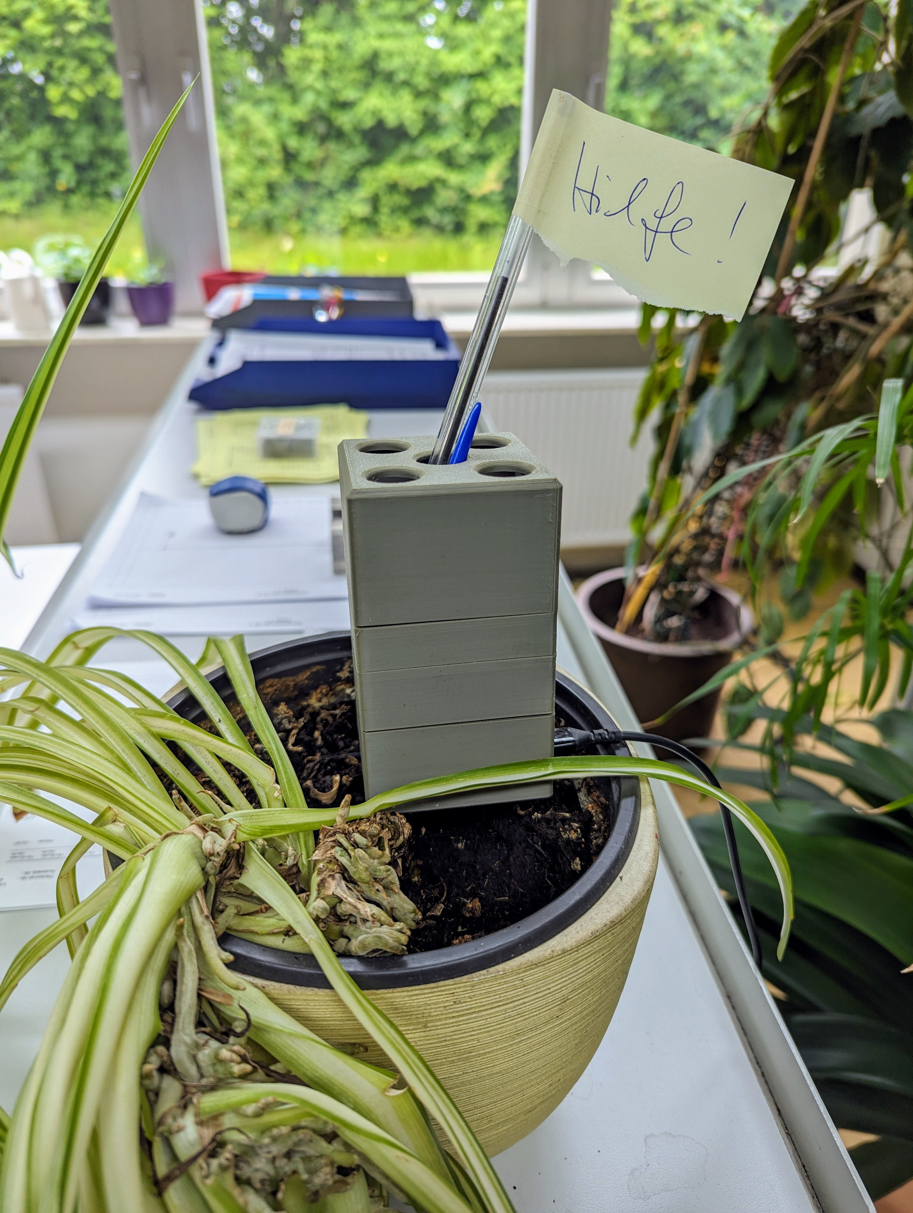 A case in the shape and size of a tea caddy with a pen protuding ouf of the top. A post-it with &ldquo;Help&rdquo; written on it sticking to the top of the pen. The case sits on the soil in a flowerpot.