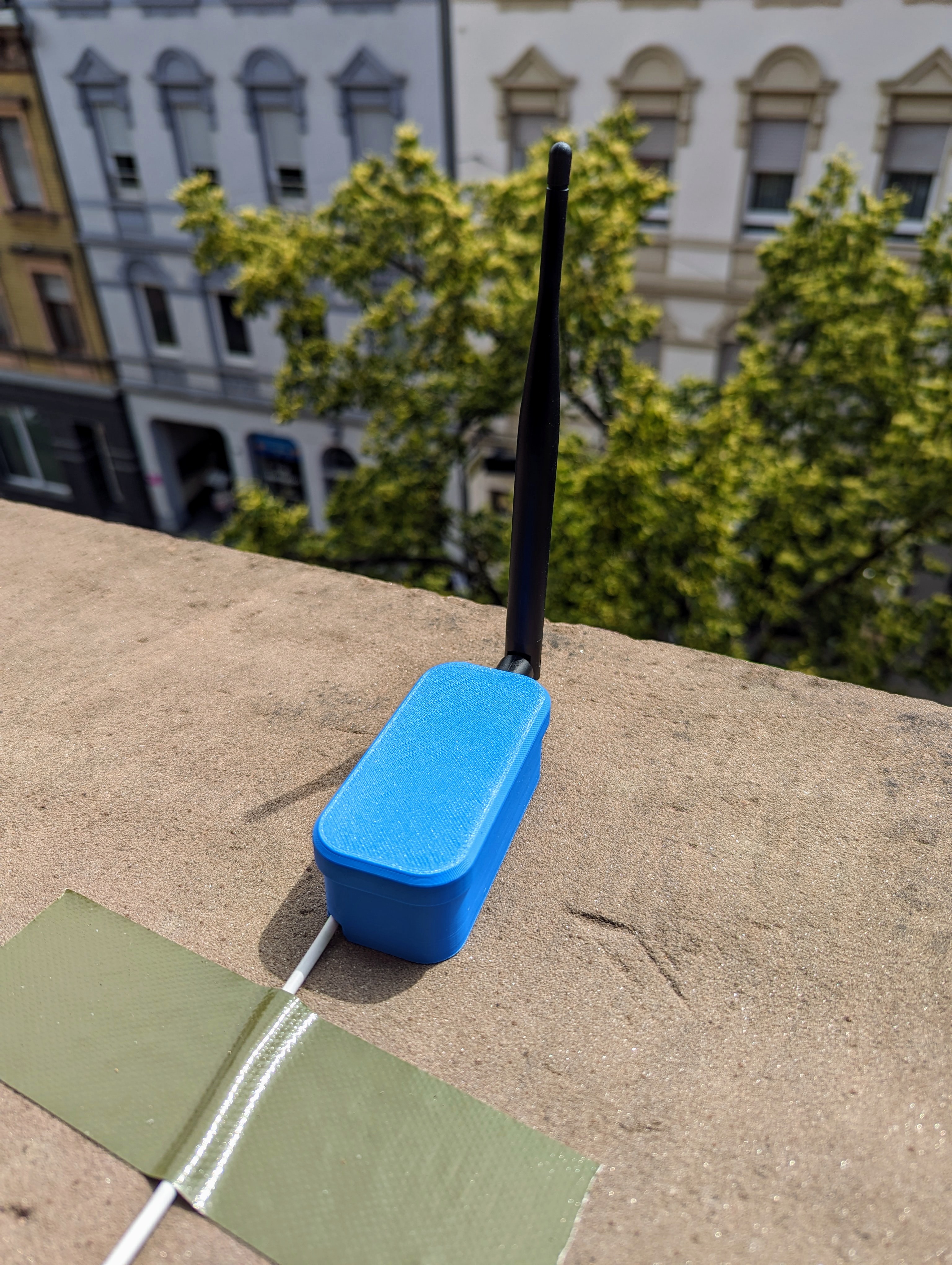 A small rectangular box with an antenna standing on an outside window sill. A usb power cable is coming out of the box, it is fixed to sill with sticky tape.